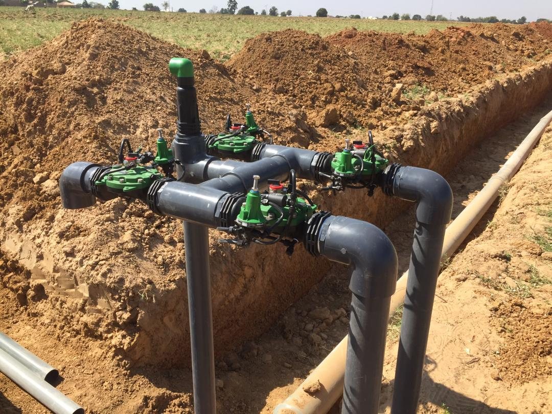 Four green hydraulic valves on a grey PVC manifold standing over an open trench.