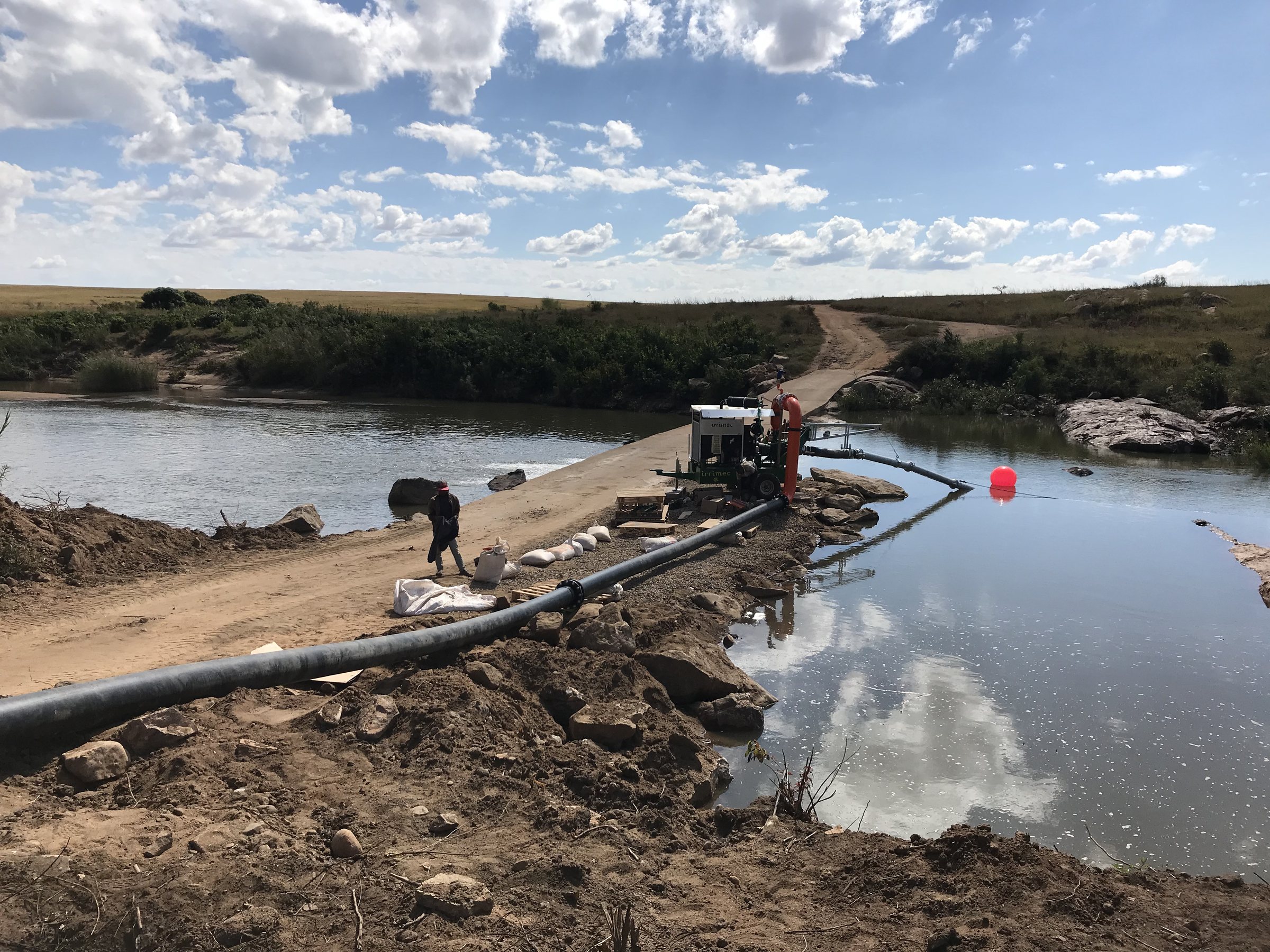 An engine-driven pump on a river causeway, its suction line running out to a red float, with the delivery pipe laid along the bank.