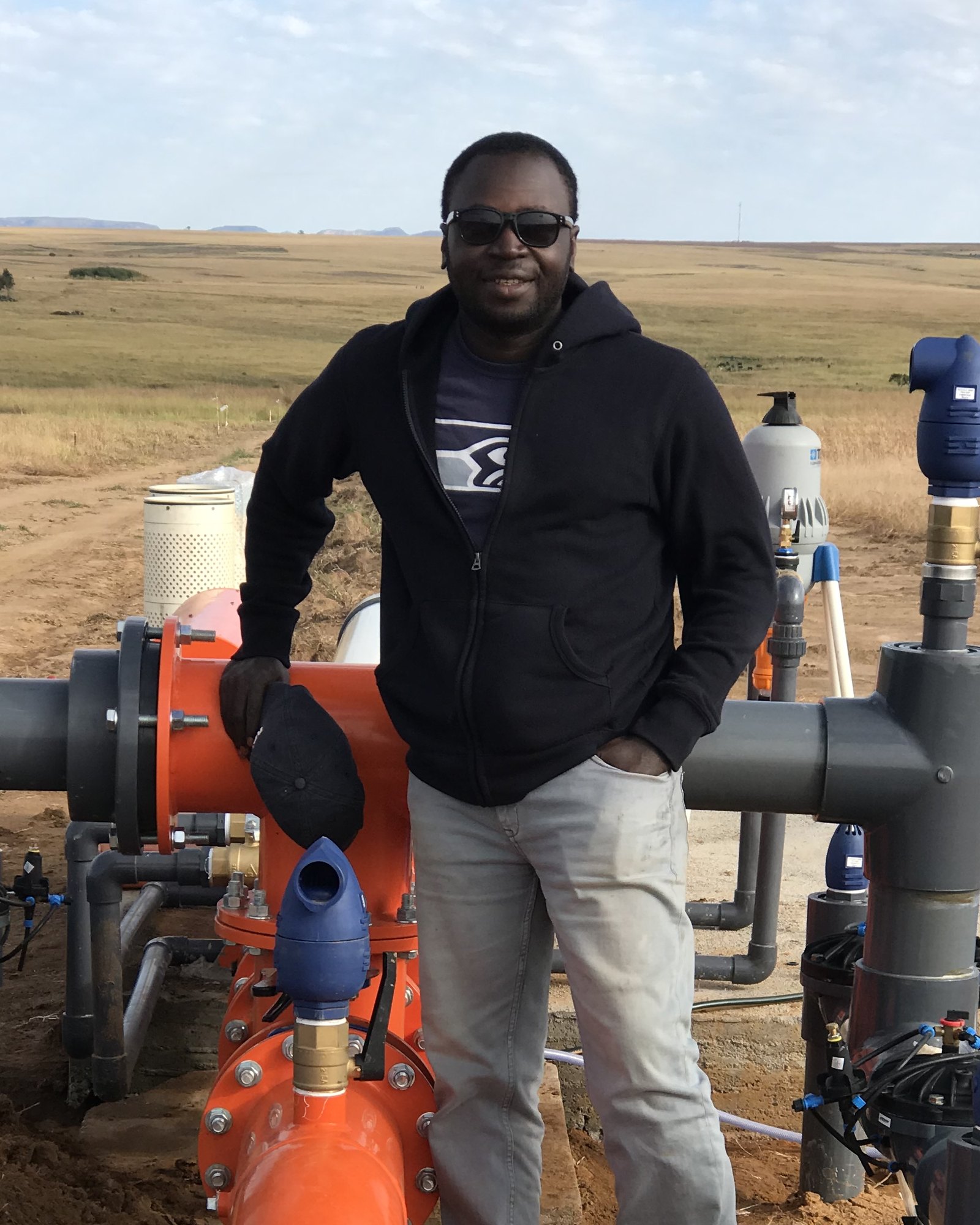 Paul Camille N’DRI standing beside the filter and valve station of an irrigation network in the field.