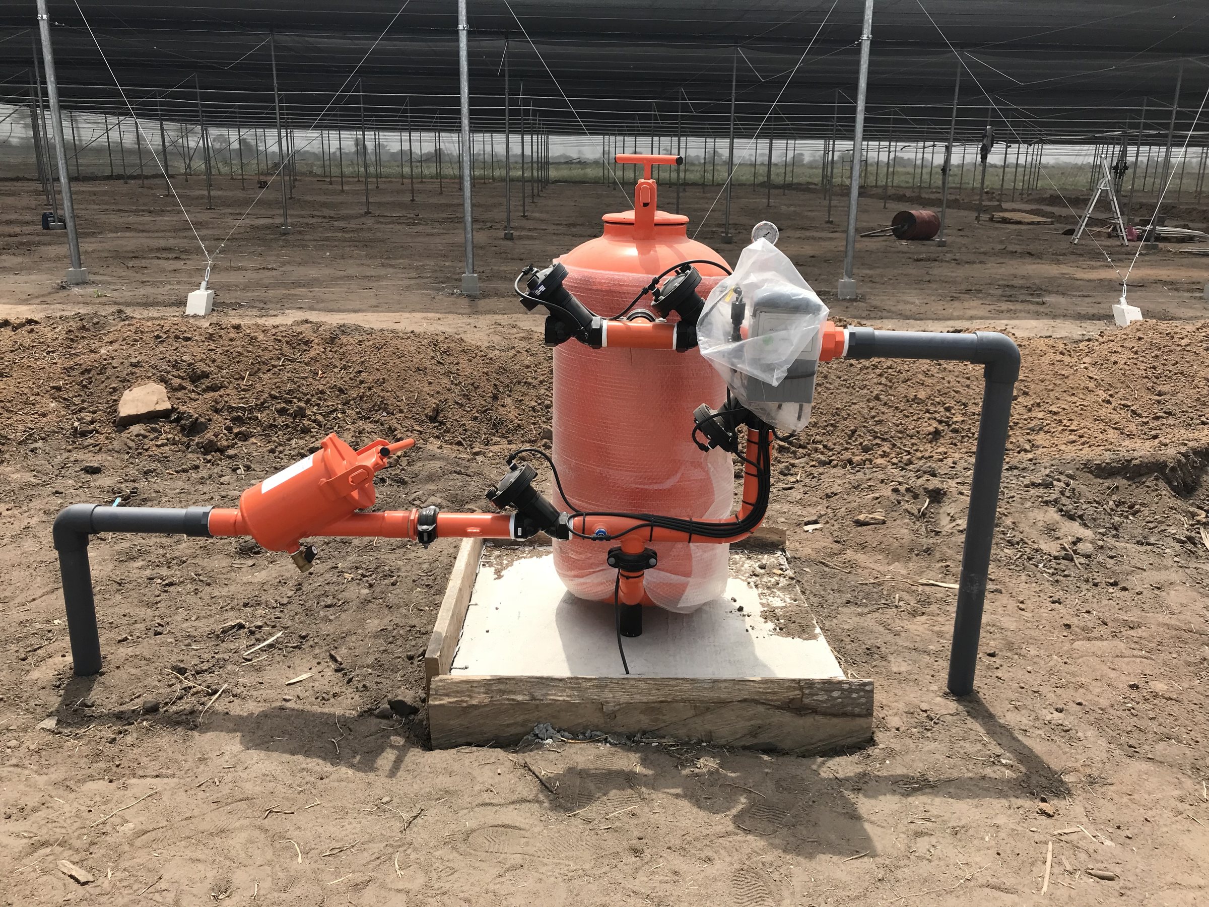 An orange fertiliser tank and a filter on a concrete plinth inside a nethouse, connected to grey PVC pipe.