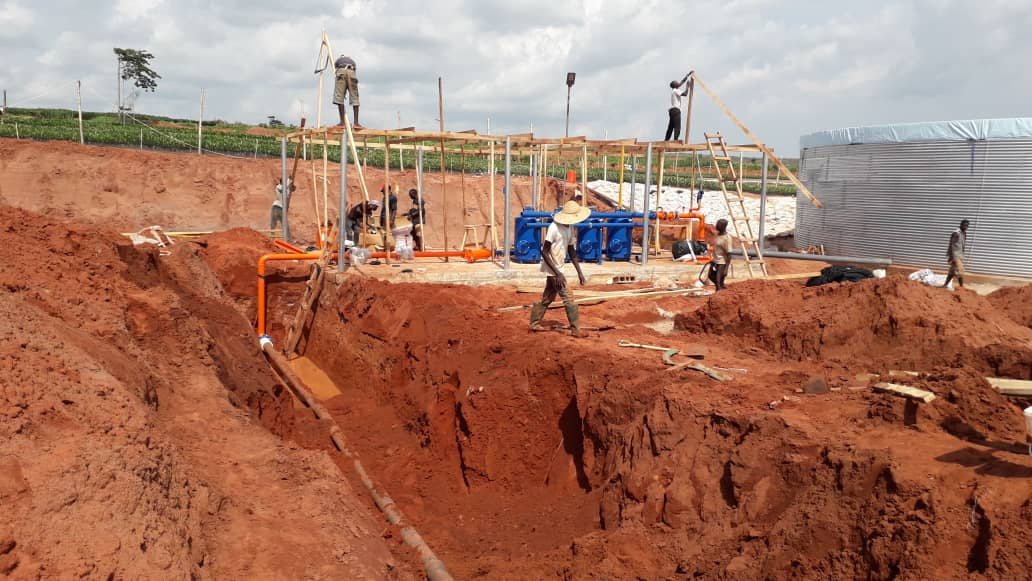 A pump house being built: a trench in red soil, workers on a timber frame, and a steel water tank to the right.